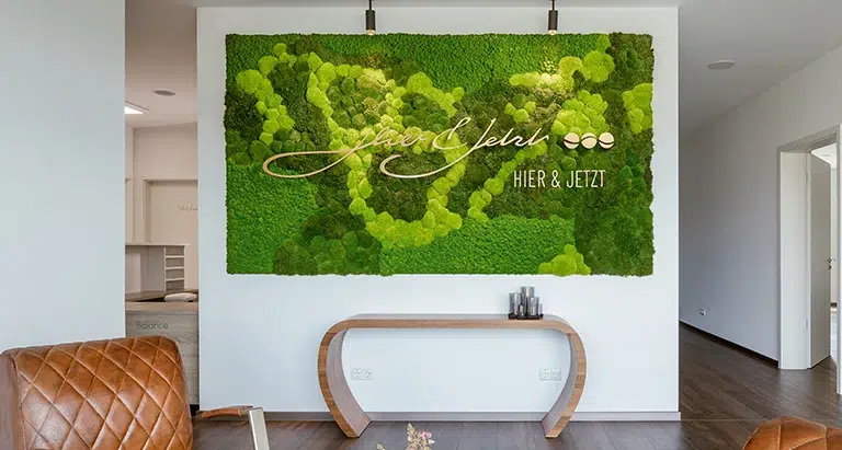 Grüne Wand aus Moos von Moosmoos im Empfangsbereich mit Schriftzug „Hier & Jetzt“, modernes Raumdesign.